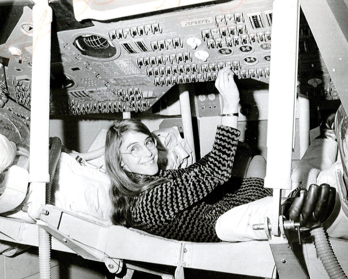 Margaret Hamilton sonriendo, tumbada en el asiento de la maqueta del módulo de mando del Apolo, con el brazo levantado hacia el panel de mandos lleno de interruptores