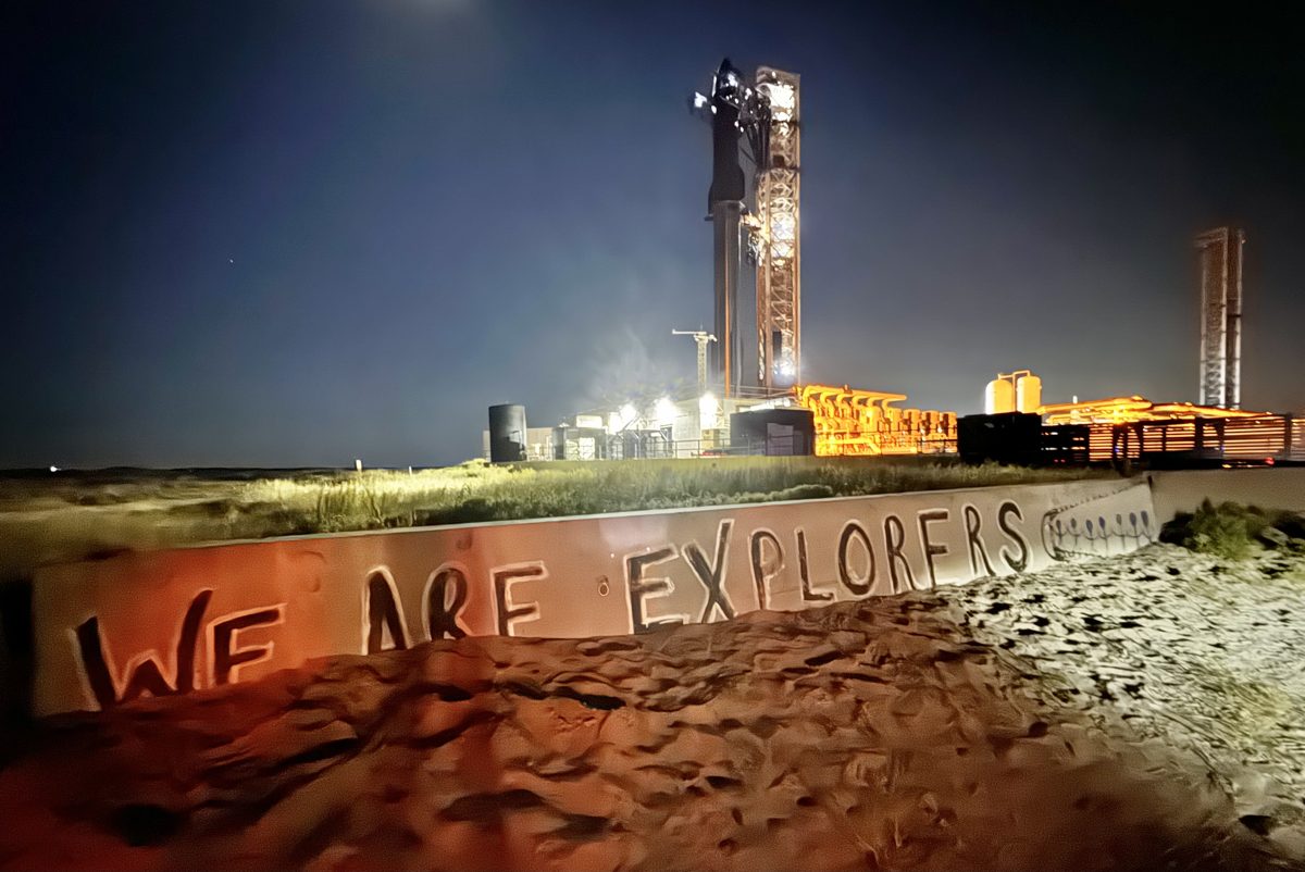 Starship sobre su plataforma de Starbase, iluminada de noche, con un muro pintado con la frase We are explorers en primer plano