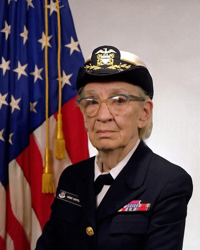 Retrato oficial de Grace Hopper con uniforme de comodoro de la Marina de Estados Unidos, junto a la bandera