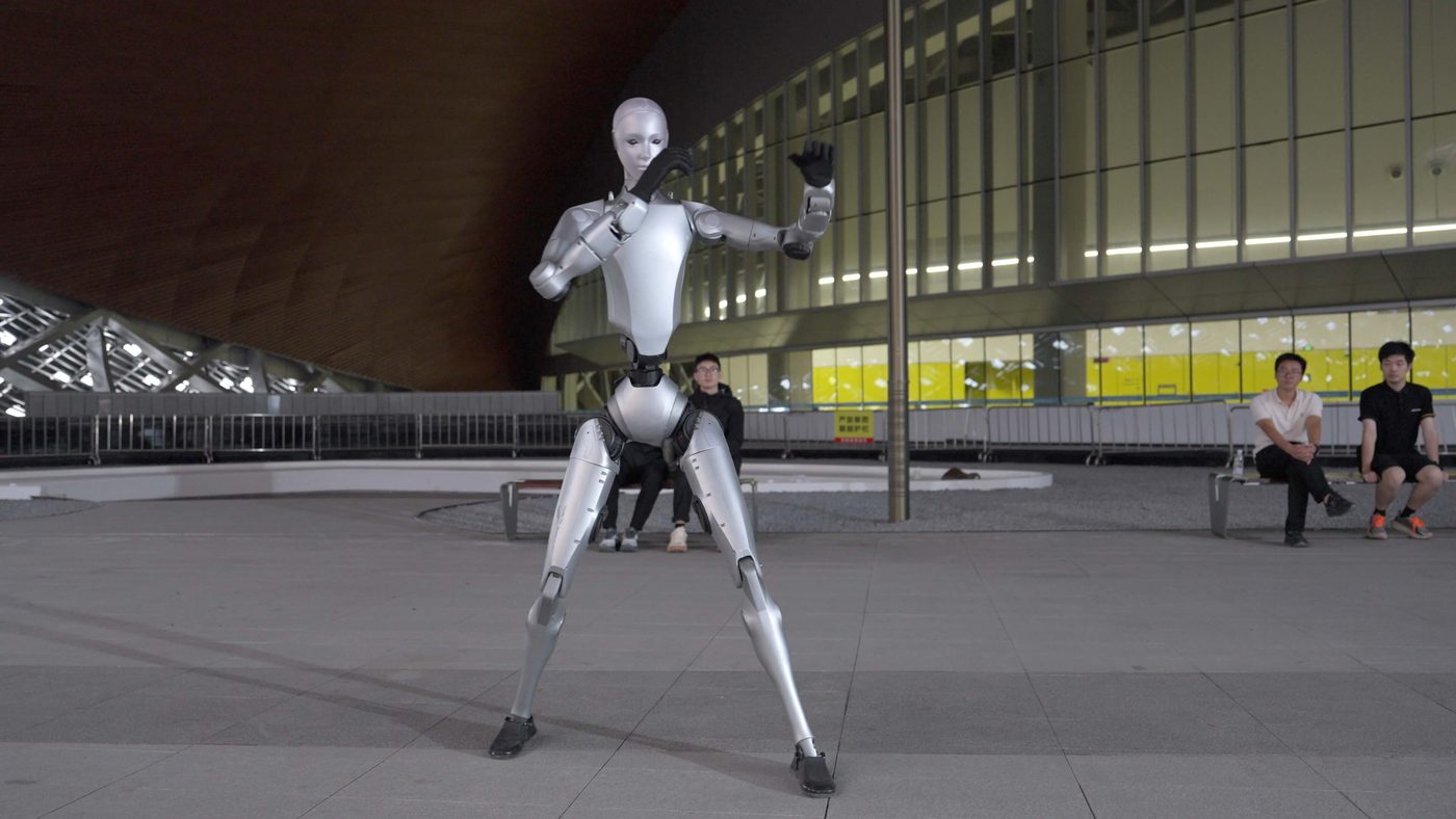 El humanoide Unitree H2 plateado en posición de kung fu en una plaza de noche, con varias personas sentadas detrás